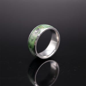 Puede incluir: Un anillo de metal plateado con una incrustación de placa de circuito verde alrededor de la banda exterior. La incrustación muestra intrincados trazos de color cobre, pequeños puntos blancos y un componente electrónico negro.