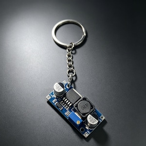 May include: A rectangular blue electronic circuit board keyring featuring two silver cylindrical capacitors, a black integrated circuit chip, a black inductor coil labelled 470, and a small blue potentiometer with a gold screw, attached to a metal ring.