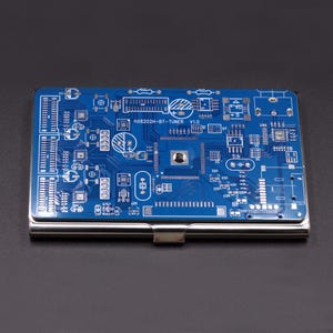 May include: A rectangular metal business card holder featuring a blue printed circuit board design on the top surface. The board displays white electronic traces, various components, and the text HX8202H-BT-TUNER V1.0 printed in white.