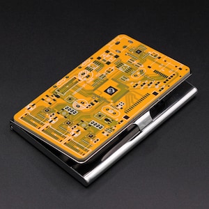 Op de afbeelding: Een rechthoekige metalen visitekaarthouder met een bovenpaneel voorzien van een geel en oranje printplaatpatroon. Het ontwerp bevat ingewikkelde elektronische sporen, componenten en de tekst HXB203H-BT-TUNER V1.0. De basis is glanzend zilver.