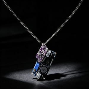 May include: A silver-toned metal chain necklace featuring a pendant made from two stacked electronic circuit boards. The larger board is blue with black components, including a circular inductor labeled 470, while the smaller board is purple.