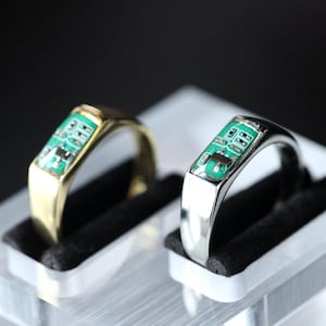 Könnte beinhalten: Zwei Ringe, einer goldfarben und einer silberfarben, jeweils mit einer rechteckigen Einlage aus einer grünen Leiterplatte, auf der kleine metallische elektronische Bauteile montiert sind. Die Ringe stehen aufrecht in einem Kunststoffhalter.