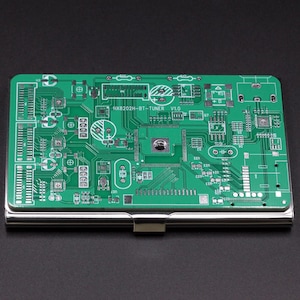 May include: A rectangular metal business card holder featuring a green printed circuit board design on the top surface. The circuit board pattern includes white lines, various electronic components, and the text HX8202H-BT-TUNER V1.0.