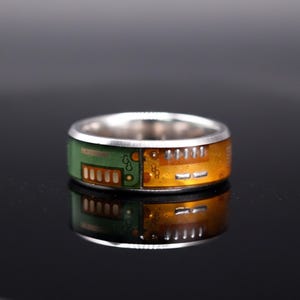 Puede incluir: Un anillo de banda en tono plateado con una incrustación de componentes de placa de circuito impreso en colores verde y ámbar. El anillo tiene un acabado metálico liso en los bordes superior e inferior, con trazas electrónicas visibles.