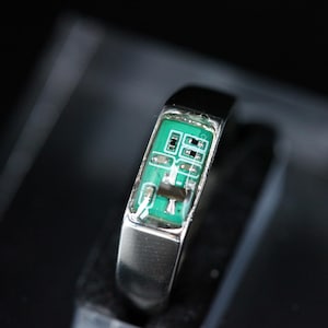 Puede incluir: Un anillo de metal plateado con una incrustación rectangular de una placa de circuito impreso verde. El componente electrónico está incrustado en la banda, mostrando pistas de cobre visibles, pequeñas resistencias soldadas y circuitos integrados.