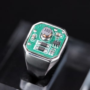 Puede incluir: Un anillo de metal plateado con una incrustación rectangular de placa de circuito verde. La placa muestra varios componentes electrónicos, incluyendo un inductor circular central marcado con el número 102 y pequeñas resistencias.