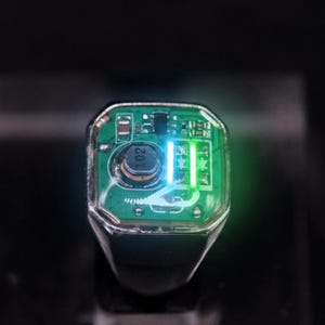 May include: A silver-toned ring featuring a green circuit board inlay under a clear protective layer. The electronic component displays a circular black inductor labeled 102 and two vertical bright green LED lights that are illuminated.