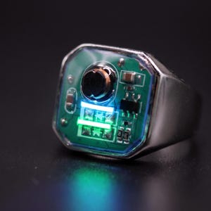 Könnte beinhalten: Ein silberfarbener Ring mit einer quadratischen Einlage aus einer grünen Leiterplatte. Die Anzeige der elektronischen Bauteile umfasst ein kleines schwarzes kreisförmiges Element, mehrere winzige Metallwiderstände und zwei leuchtende Dioden.