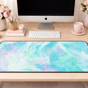 May include: A rectangular desk mat featuring a marbled pattern with swirling shades of turquoise, light blue, and soft lavender. The mat has a smooth surface and a thin black border, positioned on a light wood desk in front of a computer.