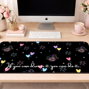 May include: A rectangular black desk mat featuring a repeating pattern of colorful balloons shaped like mouse heads, fireworks, and small stars. The text "If you can dream it, you can do it..." is printed in white cursive script across the center of the mat.