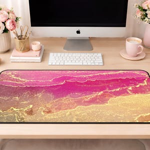 Puede incluir: Una alfombrilla de escritorio rectangular con un diseño abstracto de tinta alcohólica en tonos vibrantes de magenta, rosa intenso y burdeos profundo, acentuado por vetas doradas metálicas fluidas que crean un efecto marmolado y en capas.