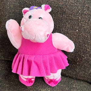 Puede incluir: Un juguete de peluche rosa con forma de hipopótamo que lleva un vestido fucsia y zapatillas de ballet. El hipopótamo tiene una flor morada en la cabeza y está saludando con el brazo izquierdo. El juguete es de felpa suave.