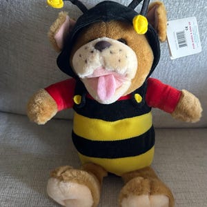 May include: A plush toy dog dressed as a bee. The dog has brown fur, a black and yellow striped body, and a black hood with antennae. It wears a red shirt and has its tongue sticking out.