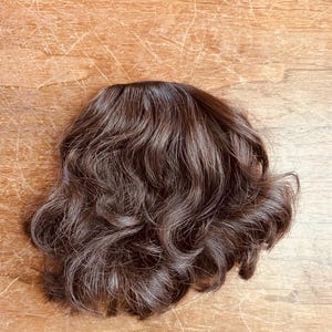 Op de afbeelding: Een donkerbruine, golvende pruik met een natuurlijke uitstraling. De pruik is gestyled met losse krullen en heeft een realistische haarlijn. Het haarstuk is ontworpen om volume en lengte toe te voegen aan het haar.