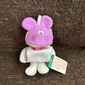Peut inclure: Un lapin en peluche avec une tête violette, un corps blanc et un nœud rose. Le lapin a des moustaches noires et une étiquette avec du texte. Idéal comme décoration ou cadeau de Pâques.