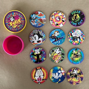 May include: A collection of Felix the Cat collectible game pieces. The pieces are round with various colourful designs featuring the cartoon cat. A pink container and a yellow and purple container with the words "Felix the Cat" are also present.