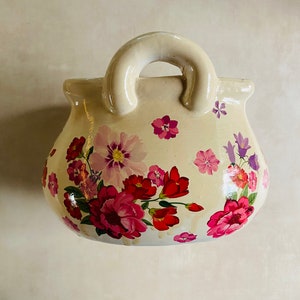 Puede incluir: Un macetero de cerámica blanca con forma de bolso de mano con un diseño floral con flores rosas, rojas y moradas. El macetero tiene un asa y es perfecto para exhibir plantas o flores pequeñas.