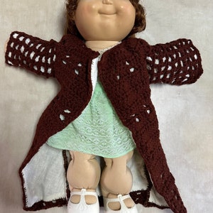 May include: A Cabbage Patch Kid doll wearing a brown crocheted coat over a green dress and white shoes.