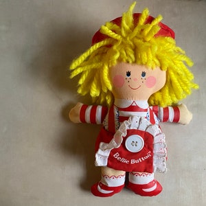 May include: A red and white striped doll with yellow yarn hair. The doll is wearing a red dress with white lace trim and a white button. The doll's dress says "Bellie Button".