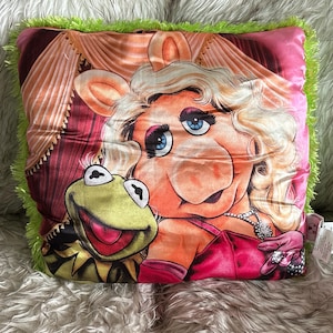 May include: Square decorative pillow featuring Miss Piggy and Kermit the Frog. The pillow has a pink satin background with a green fringe border. Miss Piggy is wearing a pink dress and jewelry. Kermit is smiling. The pillow is on a fluffy, white surface.