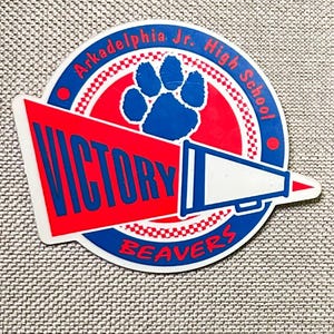 May include: A circular sticker with a red and blue design. The sticker features the text "Arkadelphia Jr. High School" and "Victory" in red and blue. A blue paw print and a megaphone are also present, with the word "Beavers" at the bottom.