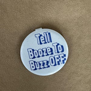 May include: A round, white button with the text "Tell Booze To Buzz OFF" in blue, bold, capital letters. The button has a small metal clasp on the top edge. The background is a solid, tan color.