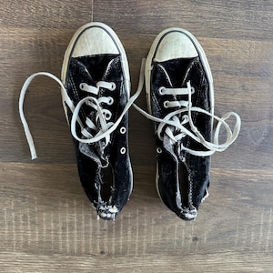 May include: Pair of worn black canvas sneakers with white laces and rubber soles. The shoes show signs of wear and tear, with frayed edges and a vintage aesthetic. The sneakers are placed on a wooden surface.