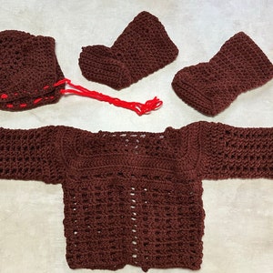 Puede incluir: Un conjunto de bebé de punto de color marrón que incluye un jersey, botines y un gorro. El jersey tiene mangas largas y un diseño sencillo. Los botines y el gorro también están tejidos con un diseño sencillo.