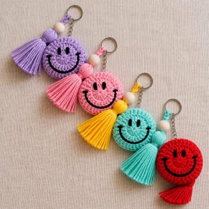 Pode incluir: Quatro porta-chaves redondos de croché com rostos sorridentes pretos bordados. Cada porta-chaves inclui uma borla de fio a condizer, uma conta de madeira e um anel de metal prateado. As cores são lavanda, rosa, amarelo e vermelho.