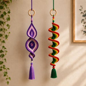 Op de afbeelding: Twee hangende gehaakte ornamenten aan houten ringen. Het linker ornament heeft paars en wit garen in een gedraaide vorm met een centrale bloem en een paarse kwast. Het rechter ornament is een spiraal van rood, geel en groen garen met een groene kwast.
