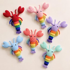 Puede incluir: Seis juguetes de langosta tejidos a ganchillo dispuestos en grupo. Cada langosta presenta una cabeza y pinzas de color sólido en rojo, rosa, morado, azul claro, coral o verde menta, junto con una cola y patas a rayas con colores del arcoíris.