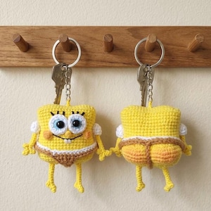 May include: Two yellow crochet keychains shaped like a cartoon character with large eyes, a wide smile, and small arms and legs. Each keychain features a metal ring with a key attached, showcasing the front and back views of the character wearing brown underwear.