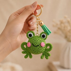 May include: A green crocheted frog keychain featuring a round body, two black eyes with white borders, pink circular cheeks, and a small embroidered smile. The frog is attached to a gold-toned metal keyring by a chain with a light green tassel.