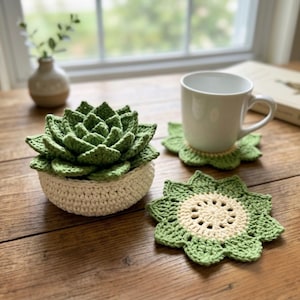 May include: A crocheted green succulent plant in a cream-colored pot, accompanied by two matching flower-shaped coasters. One coaster is placed under a white ceramic mug, while the other lies flat, showcasing a cream center and green petals.
