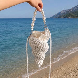 May include: A white crocheted handbag shaped like a spiral seashell, featuring a cream-colored fabric interior lining and a handle made of large, round, white faux pearls. The bag includes a long, thin, white braided cord shoulder strap.