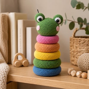 Puede incluir: Un juguete apilable de ganchillo con una cabeza de rana verde con ojos negros y mejillas rosadas, seguido de cinco anillos apilados en colores rosa, naranja, amarillo, verde y azul. El juguete está hecho de hilo con un patrón de punto visible.