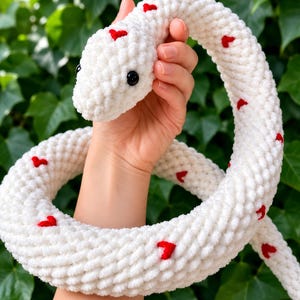 Peut inclure: Un long serpent en peluche crocheté blanc avec une texture en relief. Le serpent est orné de petits cœurs rouges dispersés sur toute sa longueur et possède deux yeux ronds en plastique noir situés sur les côtés de sa tête.
