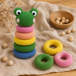 May include: A crochet stacking toy featuring a green frog head topper with black eyes and pink cheeks, sitting atop a stack of five colourful rings in pink, peach, yellow, green, and blue. Two additional rings in yellow and pink sit nearby.