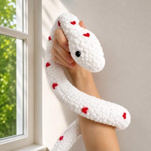 Puede incluir: Un juguete de serpiente largo, tejido a ganchillo en color blanco con un acabado de hilo suave y texturizado. La serpiente está decorada con pequeños parches rojos en forma de corazón espaciados a lo largo y tiene dos ojos negros.