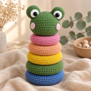 May include: A crochet stacking toy featuring a green frog head topper with black and white eyes and pink cheeks. The base consists of four stacked rings in pink, orange, yellow, green, and blue colours, all crafted with a textured yarn stitch pattern.