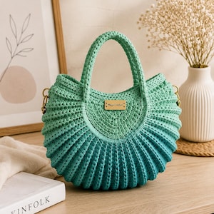 Puede incluir: Un bolso de mano en forma de media luna hecho de hilo de ganchillo con un degradado de color verde menta a azul verdoso intenso. El bolso cuenta con dos asas integradas y una pequeña etiqueta rectangular de madera con el texto Maya Crochet.