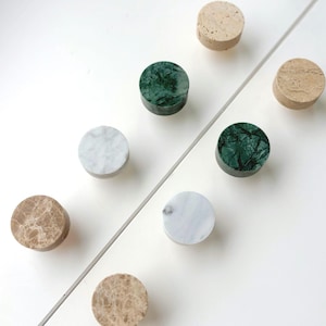 Puede incluir: Una colección de siete pomos circulares para armarios hechos de diversas piedras naturales. El conjunto incluye dos pomos de mármol verde oscuro, dos de mármol blanco con vetas grises, dos de piedra marrón texturizada y uno de piedra beige.