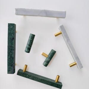 May include: A collection of cabinet hardware including rectangular bar pulls and cylindrical knobs made of white and dark green marble. Each piece features polished gold-tone metal mounting posts, showcasing natural stone veining and textures.