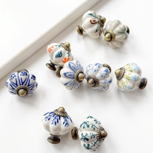 May include: A collection of eight ceramic drawer knobs with a fluted, pumpkin-like shape. Each knob features a white base with hand-painted floral patterns in blue, green, and orange, finished with a metallic bronze-toned circular base and centre.