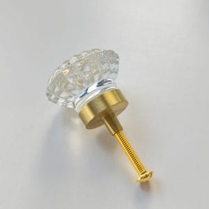 May include: A clear, faceted glass cabinet knob with a round, textured top. The base and the attached threaded bolt are made of a brushed brass-colored metal, providing a contrast to the transparent glass head of the hardware piece.