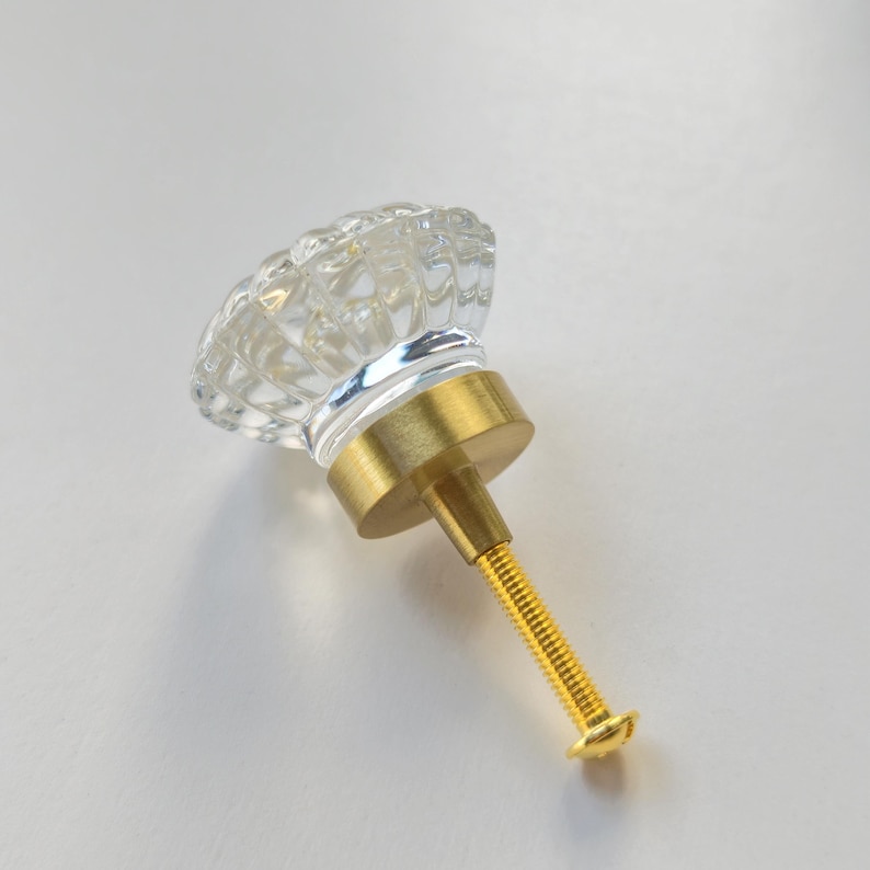 May include: A clear, faceted glass cabinet knob with a round, textured top. The base and the attached threaded bolt are made of a brushed brass-colored metal, providing a contrast to the transparent glass head of the hardware piece.