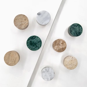 May include: A collection of seven circular knobs made from various natural stones. The set includes two white marble knobs with grey veining, two dark green marble knobs, two tan brown marble knobs, and one light beige textured stone knob.