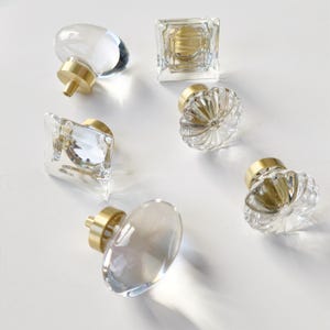 May include: A collection of six cabinet knobs featuring clear glass or crystal tops in various shapes including round, square, and fluted designs, each attached to a cylindrical brushed brass metal base for mounting to furniture or doors.