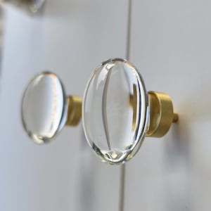 May include: Two oval-shaped cabinet knobs featuring clear, smooth glass fronts attached to brushed brass-toned cylindrical bases. The knobs are mounted on a white surface, showcasing a reflective, transparent design with metallic accents.