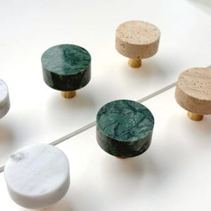 May include: A collection of circular cabinet knobs featuring flat, round tops in various stone textures including dark green marble, white marble, and beige travertine, each mounted on a small, cylindrical gold-toned metal base.